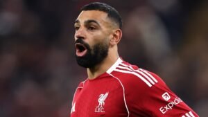 Mohamed Salah dans un maillot rouge vif de Liverpool, criant passionnément pendant un match, avec un arrière-plan de stade flou.