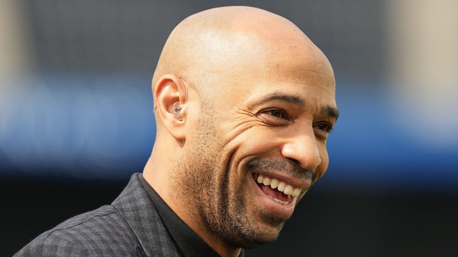 Une légende du football souriante, au crâne rasé, vêtue d'une veste noire, se détachant sur un fond de stade flou, dégageant chaleur et enthousiasme.