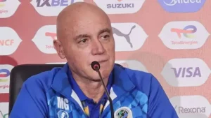 Miguel Gamondi in a blue coaching jacket, speaking at a press conference with a branded backdrop featuring various sponsor logos.