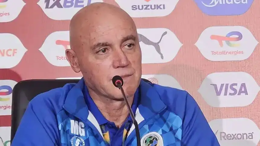 Miguel Gamondi in a blue coaching jacket, speaking at a press conference with a branded backdrop featuring various sponsor logos.