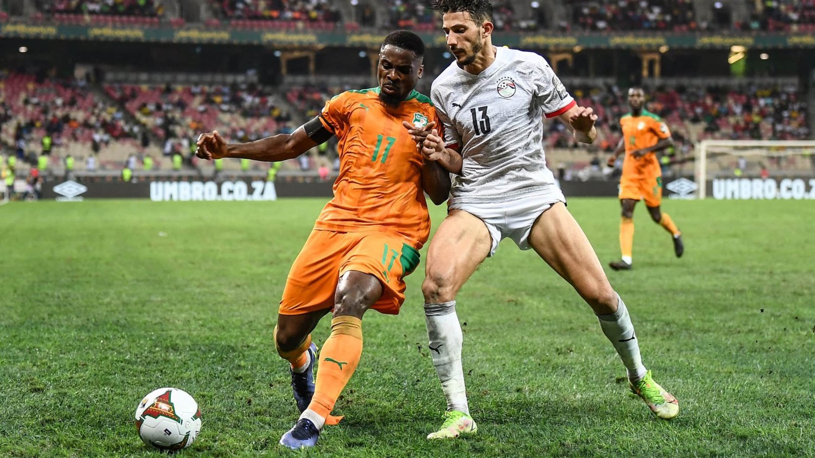 Ivory Coast’s player in orange battles for the ball with Egypt’s player in white during a tense AFCON match, with fans in the background.