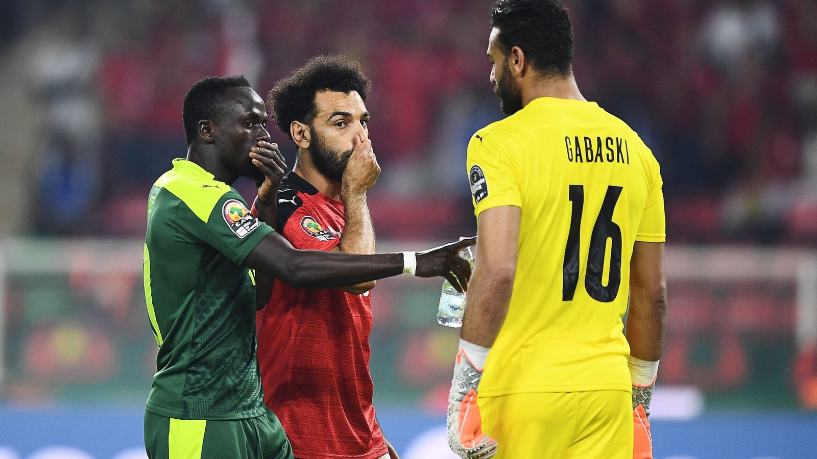 Sadio Mané et Mohamed Salah conversent de près, tandis que Gabaski, portant un maillot jaune vif, regarde lors d'un match intense de la CAN.