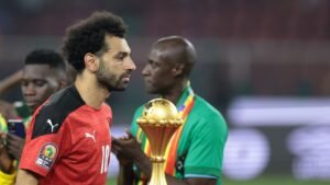 Mohamed Salah, dans le maillot rouge de l'Égypte, passe devant un coéquipier tenant le trophée de la CAN, capturant un moment de réflexion après le match.