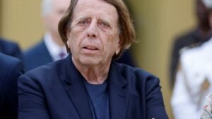 A man with thinning hair and light blue eyes wears a dark blazer, appearing contemplative while standing among a group of people.