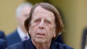 Un homme âgé pensif, aux cheveux ébouriffés, vêtu de noir, fixant intensément, sur fond flou de silhouettes indistinctes.