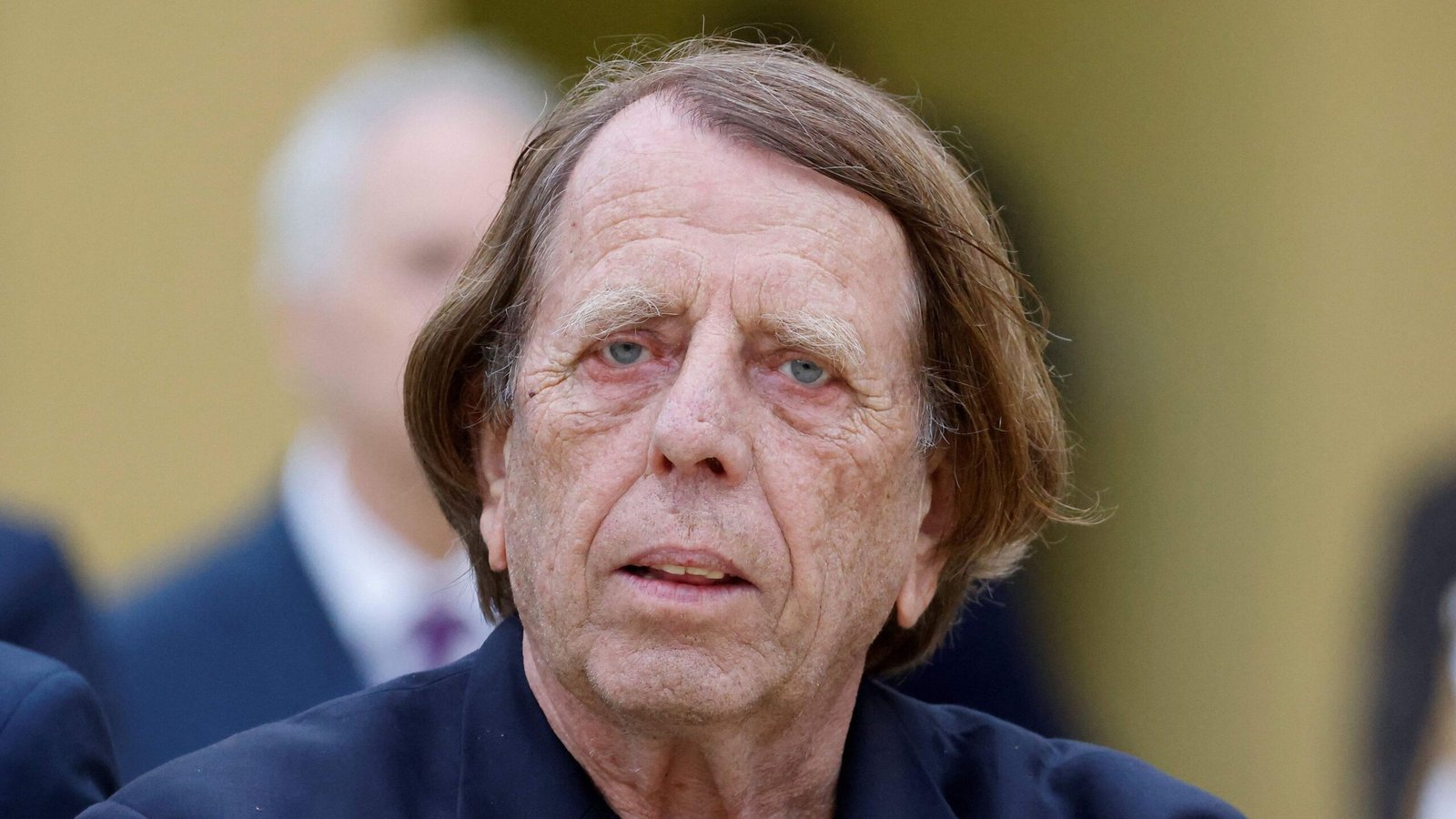 Un homme âgé pensif, aux cheveux ébouriffés, vêtu de noir, fixant intensément, sur fond flou de silhouettes indistinctes.