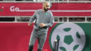 مدرب المغرب ببدلة بوما الرياضية الرمادية، يشير بإيماءات على أرض الملعب، مع أعلام المغرب وشعار الفريق واضحين في الخلفية.