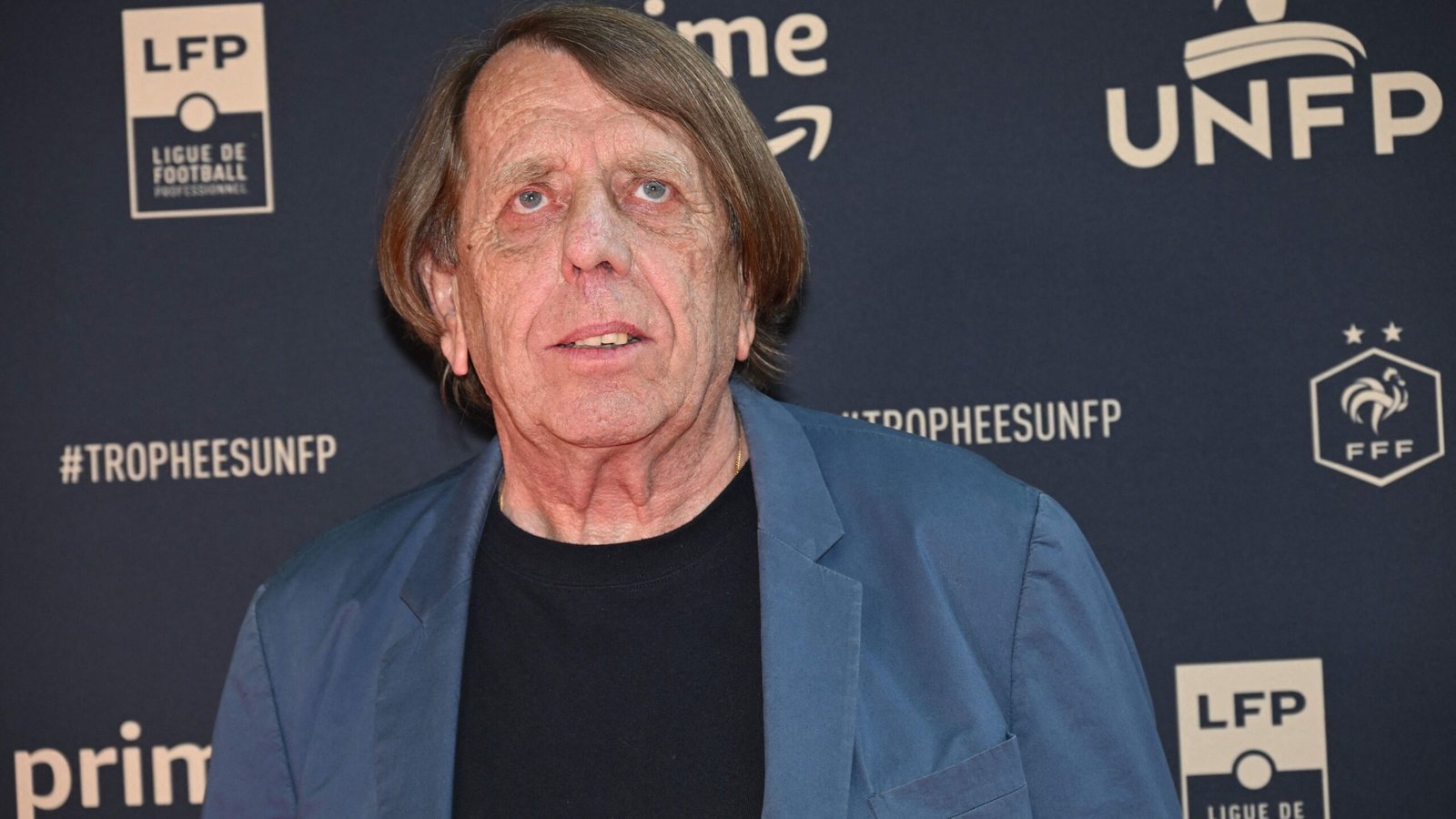 Un homme aux cheveux mi-longs se tenant devant un fond sombre arborant les logos de la LFP, de l'UNFP et de la FFF, représentant le football français.