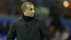 Entraîneur de football marocain en manteau noir, concentré sur le match, avec un fond de stade flou.