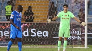Yassine Bounou in green kit gestureing during a match, with a player in blue behind him. Stadium backdrop showcases a vibrant atmosphere.