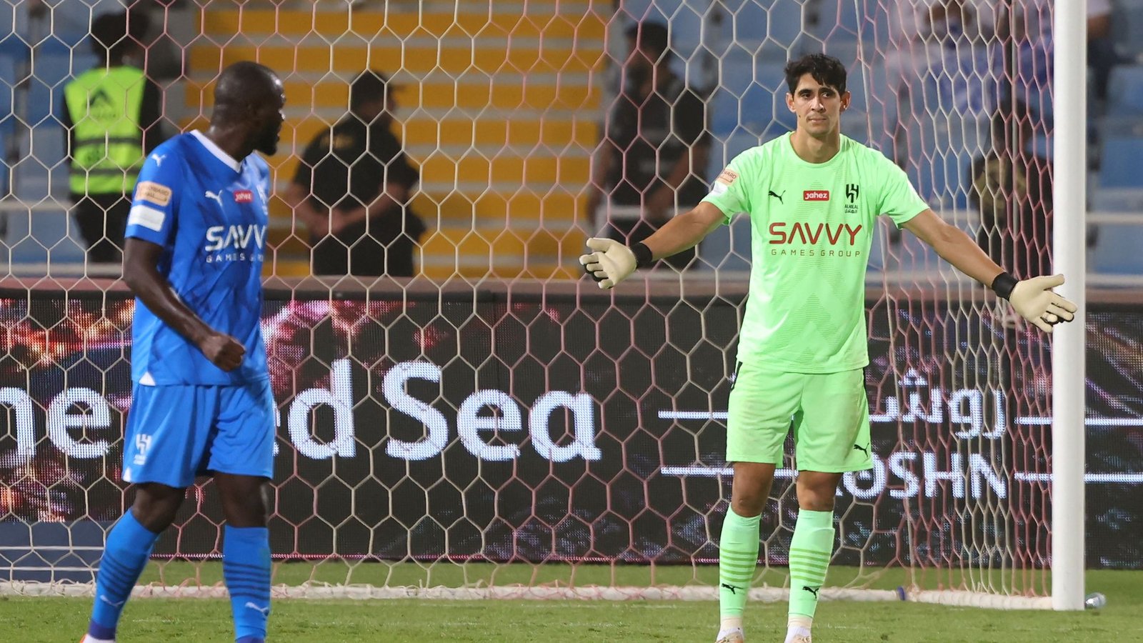 Yassine Bounou in green kit gestureing during a match, with a player in blue behind him. Stadium backdrop showcases a vibrant atmosphere.