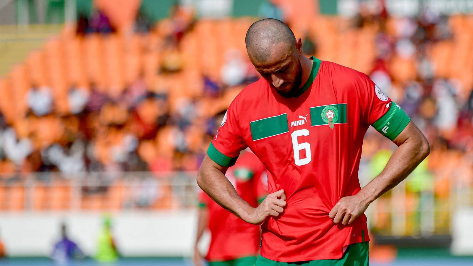Joueur marocain en tenue rouge avec des accents verts, regardant vers le bas dans un moment de déception pendant un match, avec des sièges de stade orange en arrière-plan.