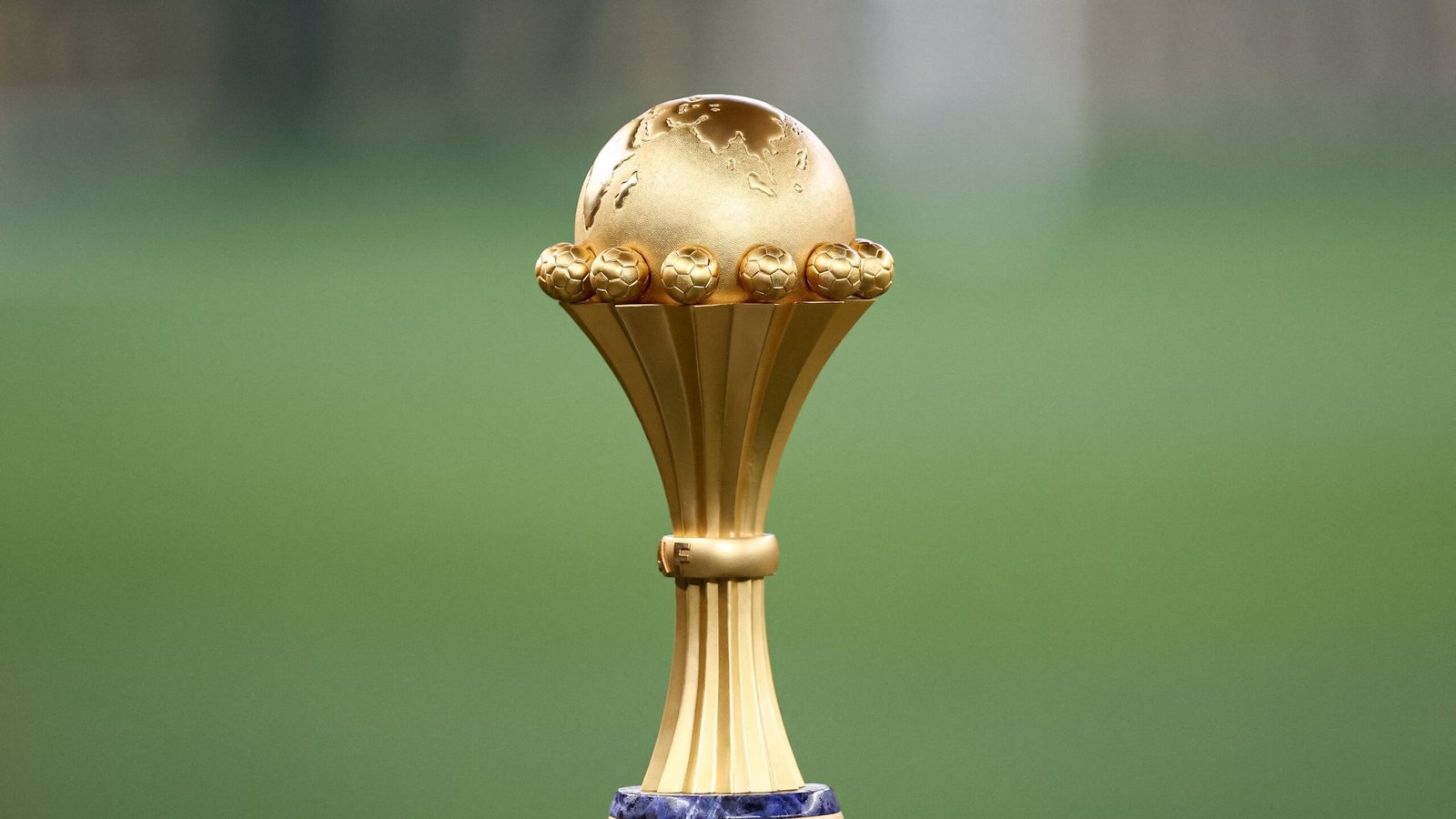 Golden Africa Cup of Nations trophy with football motifs, set against a blurred green pitch background, symbolising continental football glory.