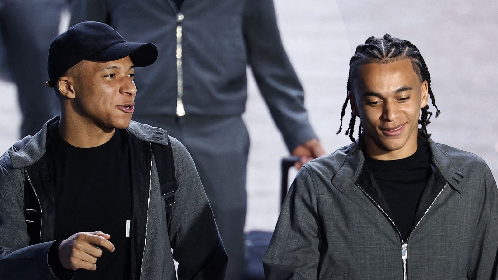 Mbappé et un coéquipier partagent un moment léger en marchant, tous deux vêtus de tenues décontractées grises, montrant une atmosphère détendue avant le match.
