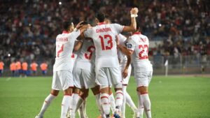 Tunisia players celebrate a goal, wearing white kits with red accents, amidst a packed stadium, showcasing team spirit and excitement.
