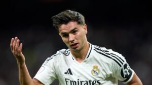 Brahim Díaz du Real Madrid exprime sa frustration par un geste pendant un match, portant un maillot blanc avec des accents noirs, sur fond de stade flou.