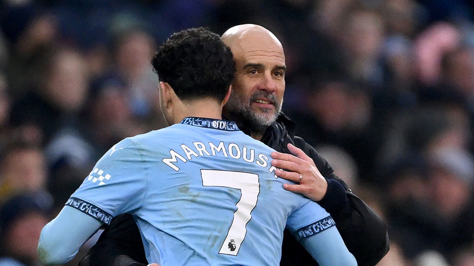 Marmoush embraces Manchester City manager Pep Guardiola, both displaying emotions post-match in a blue stadium backdrop.