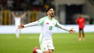 Joueur algérien célébrant un but, vêtu d'un maillot blanc avec des accents verts, lors d'un match officiel. L'excitation remplit le stade.