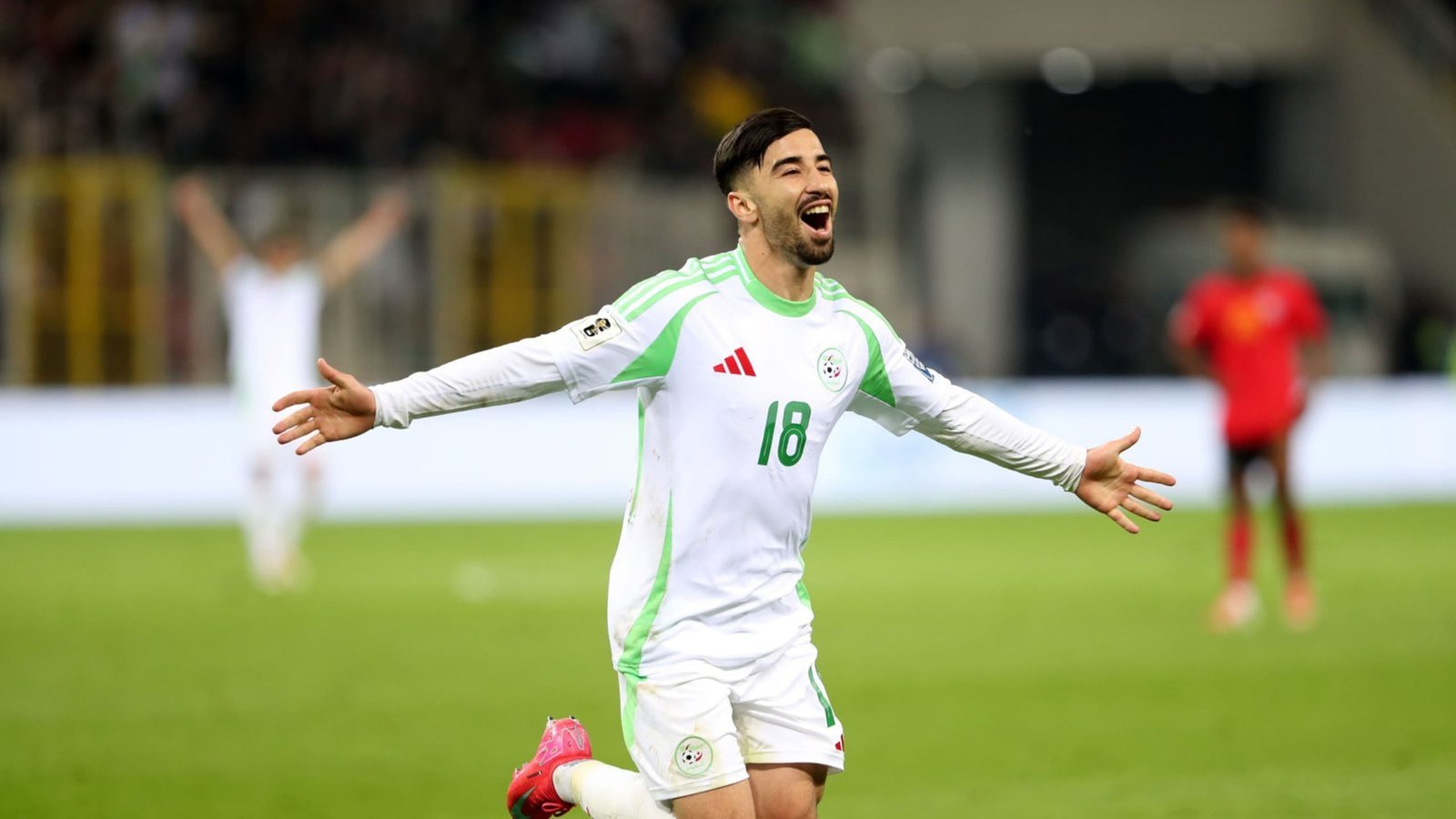 Joueur algérien célébrant un but, vêtu d'un maillot blanc avec des accents verts, lors d'un match officiel. L'excitation remplit le stade.