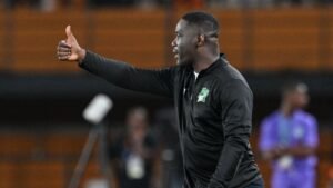 L'entraîneur ivoirien fait un geste de pouce levé pendant un match, portant un survêtement noir avec un emblème vert et blanc, sur fond de stade flou.