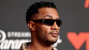 لاعب كرة قدم مغربي بشعر قصير مجعد ونظارات شمسية، يرتدي قميصًا منقوشًا، يلتقط صورة بثقة أمام خلفية مضاءة بشكل خافت.