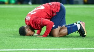 Lille player kisses the pitch in celebration after scoring, wearing a red jersey and blue shorts on a vibrant green field.