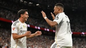 Real Madrid players celebrating a goal, wearing their white kits with black accents, against a vibrant stadium backdrop filled with fans.