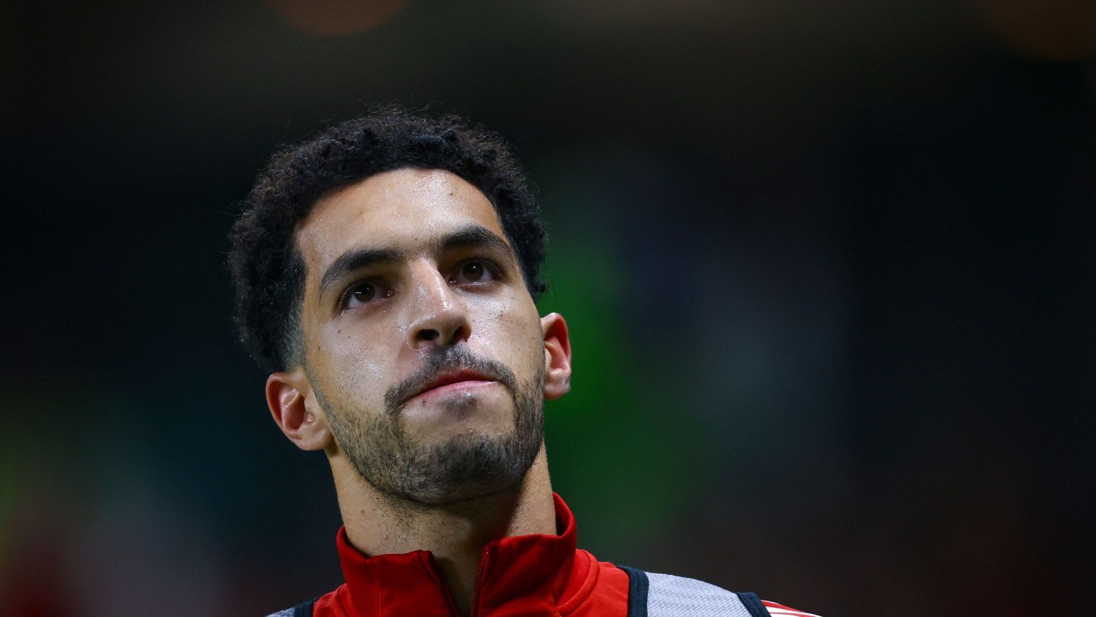 Moroccan footballer looks contemplative, wearing a red training kit, with stadium lights softly illuminating his face against a blurred background.