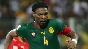 Un footballeur camerounais en maillot vert et rouge, numéro 4, sprinte avec intensité pendant un match, montrant la fierté de l'équipe.