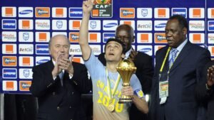 Un joueur soulève le trophée de la Coupe d'Afrique des Nations en célébration, flanqué d'officiels lors de la cérémonie de remise des prix, avec un arrière-plan vibrant.