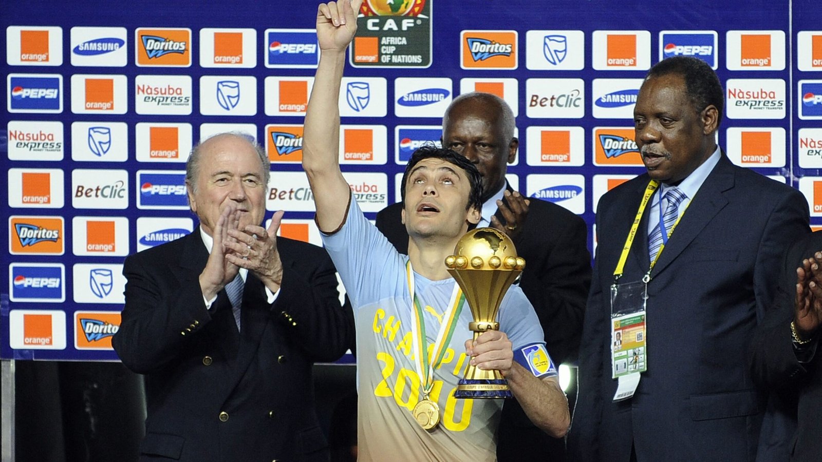 Un joueur soulève le trophée de la Coupe d'Afrique des Nations en célébration, flanqué d'officiels lors de la cérémonie de remise des prix, avec un arrière-plan vibrant.