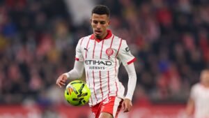 Moroccan player in a white and red Girona kit, skillfully controlling a vibrant football during a match, with fans blurred in the background.
