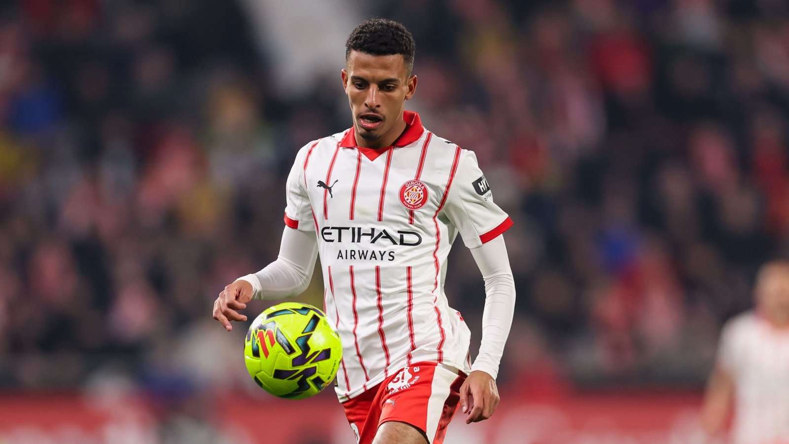 Moroccan player in a white and red Girona kit, skillfully controlling a vibrant football during a match, with fans blurred in the background.