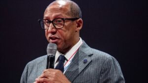 Voetbalbestuurder spreekt in een microfoon, draagt een bril en een grijs pak, tegen een donkere achtergrond.