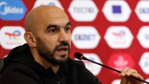 Un entraîneur de football marocain s'exprimant lors d'une conférence de presse avec un fond de Bladna News Maroc.