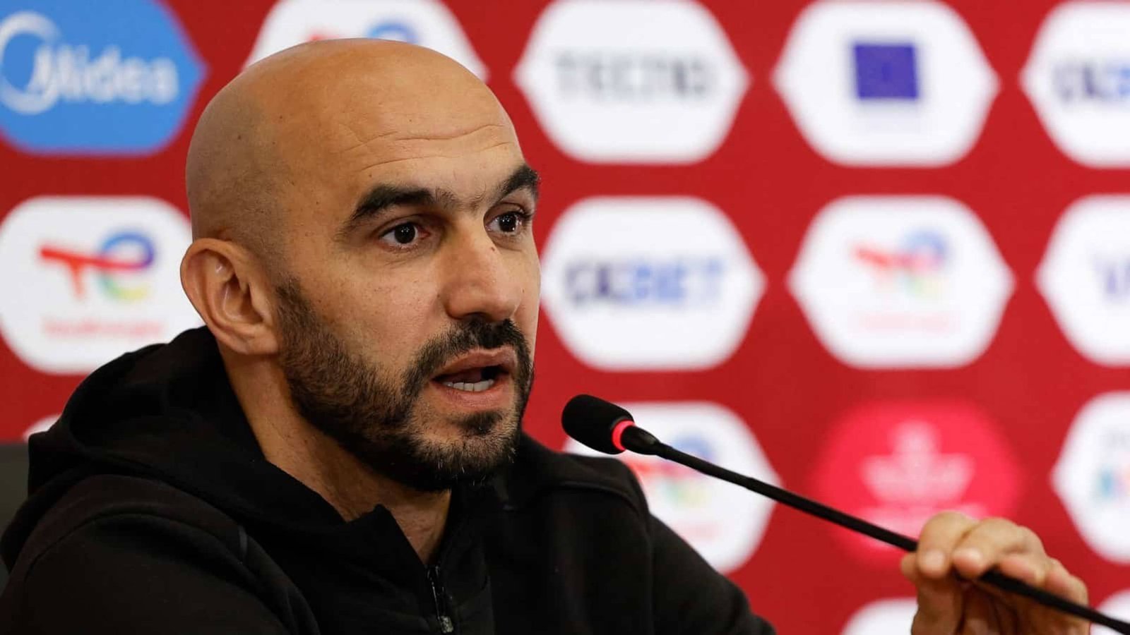 Un entraîneur de football marocain s'exprimant lors d'une conférence de presse avec un fond de Bladna News Maroc.