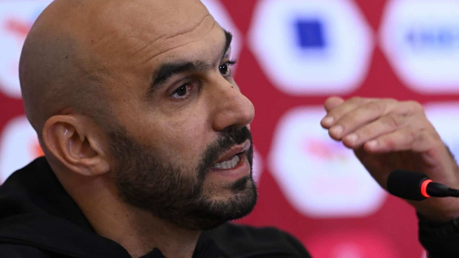 Walid Regragui fait des gestes lors d'une conférence de presse, discutant du football marocain devant un fond rouge vif orné de logos.