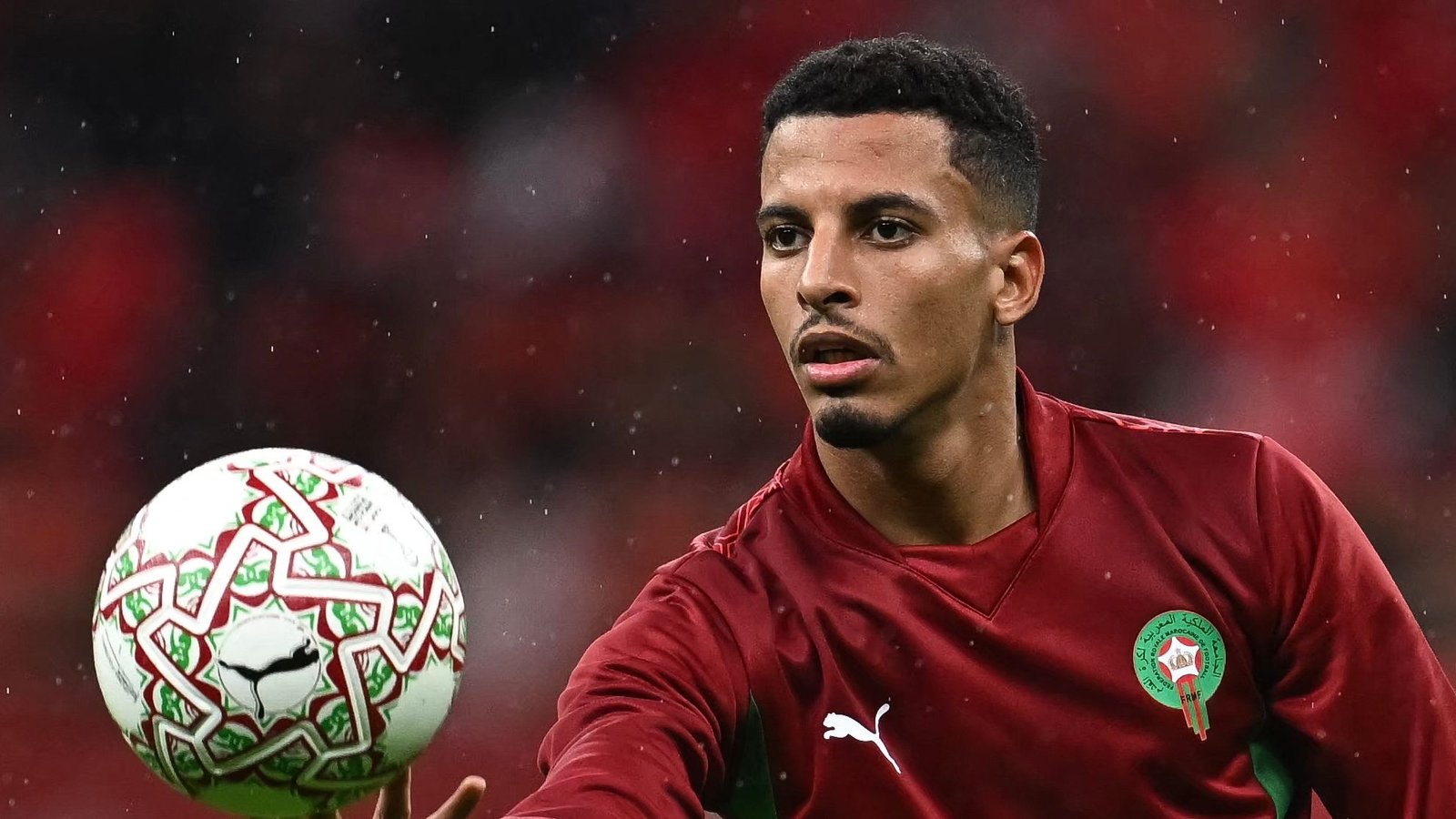 Moroccan player in maroon kit prepares to pass a decorative ball during a match, with rain droplets visible in the background.