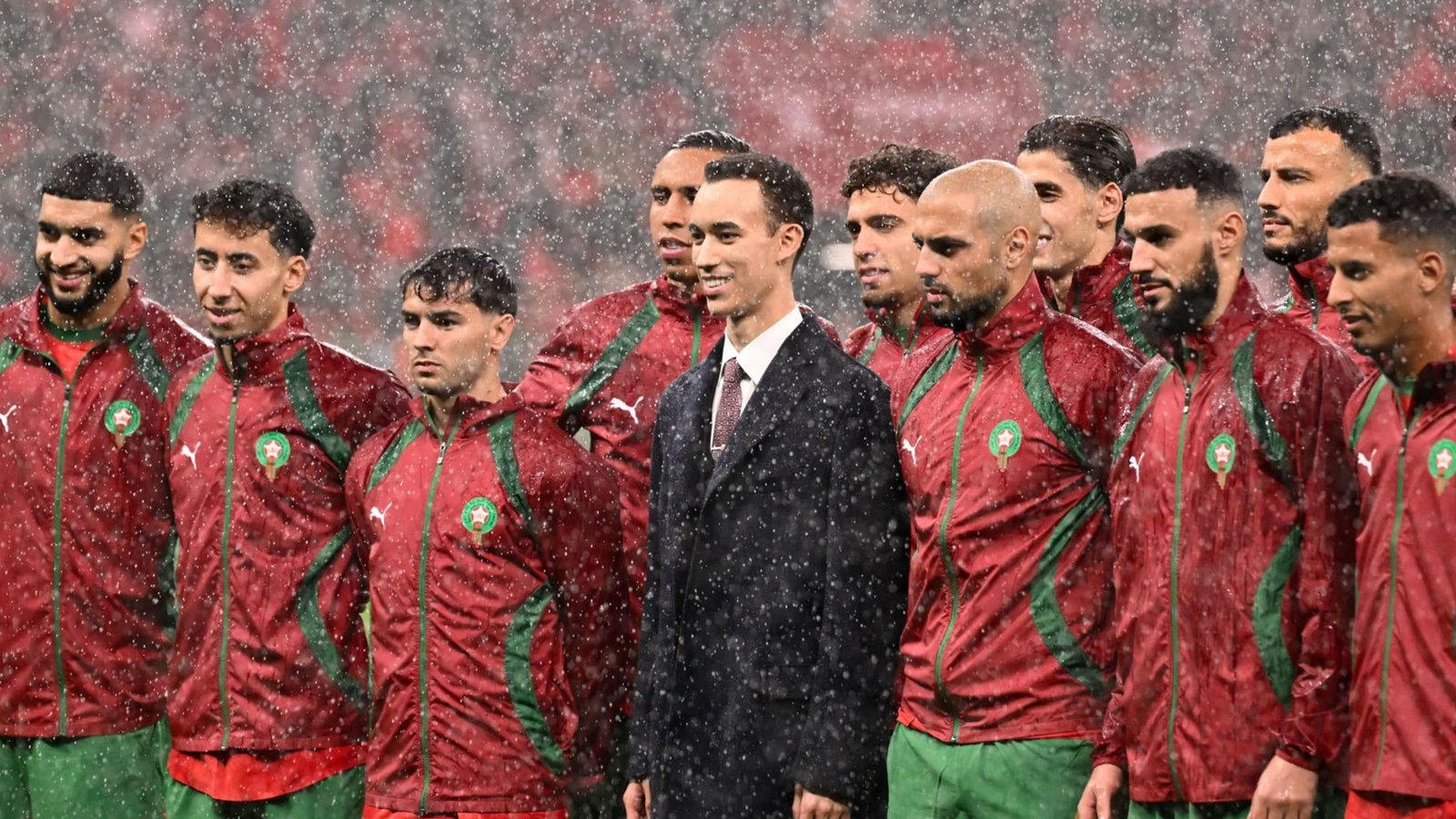 Morocco's football team stands together under rain, wearing red jackets with green accents, showcasing unity and determination pre-match.