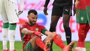 Moroccan player seated on the pitch, grimacing in pain, while teammates in red and black gear offer support during a tense moment.