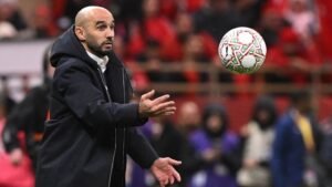 Walid Regragui gebaart naar het veld, zich voorbereidend om een voetbal te ontvangen versierd met Marokkaanse kleuren, temidden van een zee van rode fans.