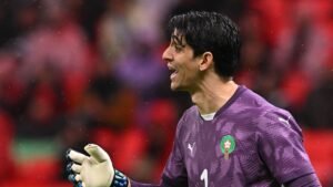 Moroccan goalkeeper yelling passionately during a match, wearing a purple kit with the national emblem, against a blurred stadium backdrop.