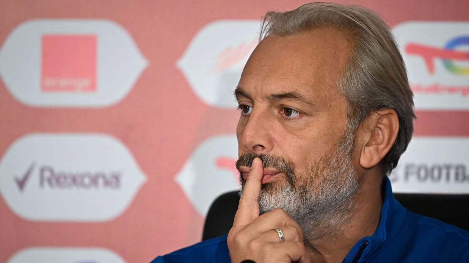 Coach with grey hair and beard gestures thoughtfully during a press conference, set against a pink background adorned with logos.