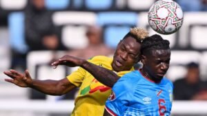 لاعب مالي يتنافس على كرة رأسية مع منافس كونغولي خلال مباراة شديدة في كأس أمم أفريقيا. ألوان الزي الزاهية والتركيز على الحركة.
