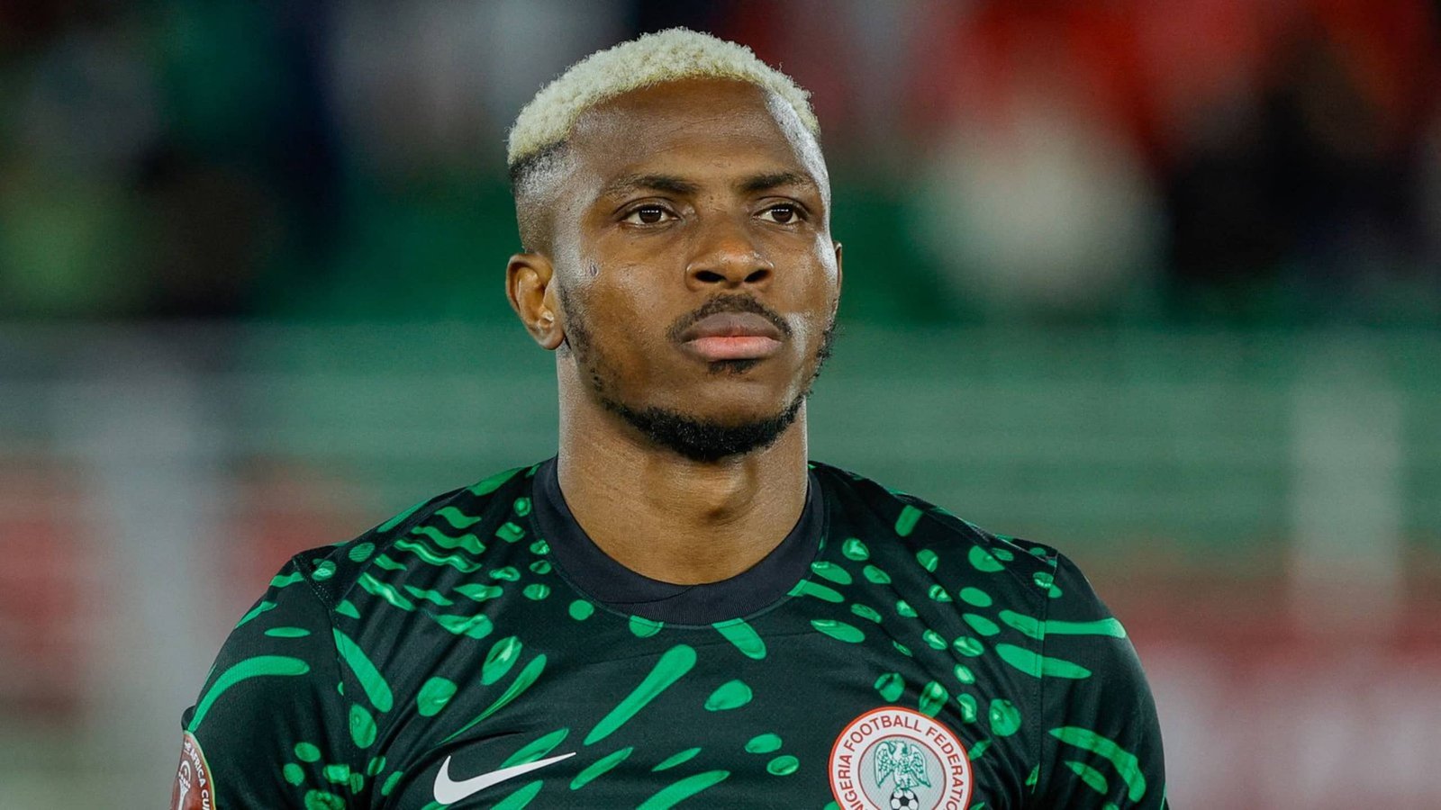 Nigeria goalkeeper in a striking green-patterned kit, gazing intently ahead with a focused expression, set against a blurred stadium backdrop.