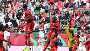 Morocco's players in red compete for a header against Algeria, as fans wave flags in the background during a tense AFCON match.