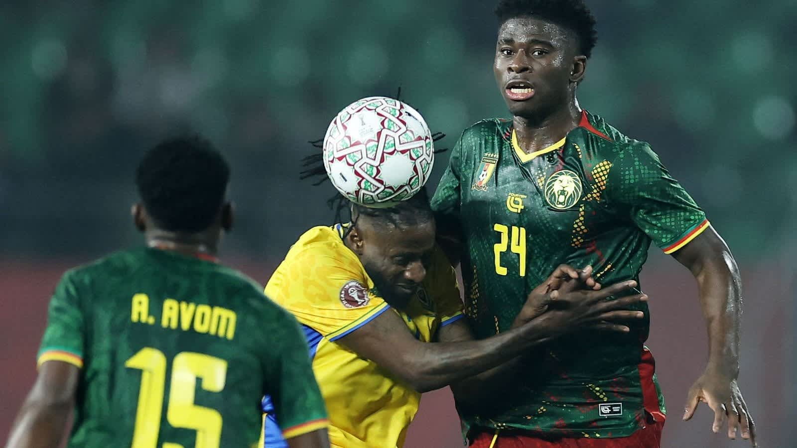 Des joueurs camerounais s'affrontent alors que l'un d'eux effectue une tête ; A. Avom (15) défend, tandis qu'un autre joueur (24) réagit lors d'un moment intense du match.