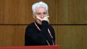 Entraîneur pensif, profondément absorbé dans ses réflexions, portant une chemise noire avec un cordon, devant un fond en bois. Traduit une concentration sur le football marocain.