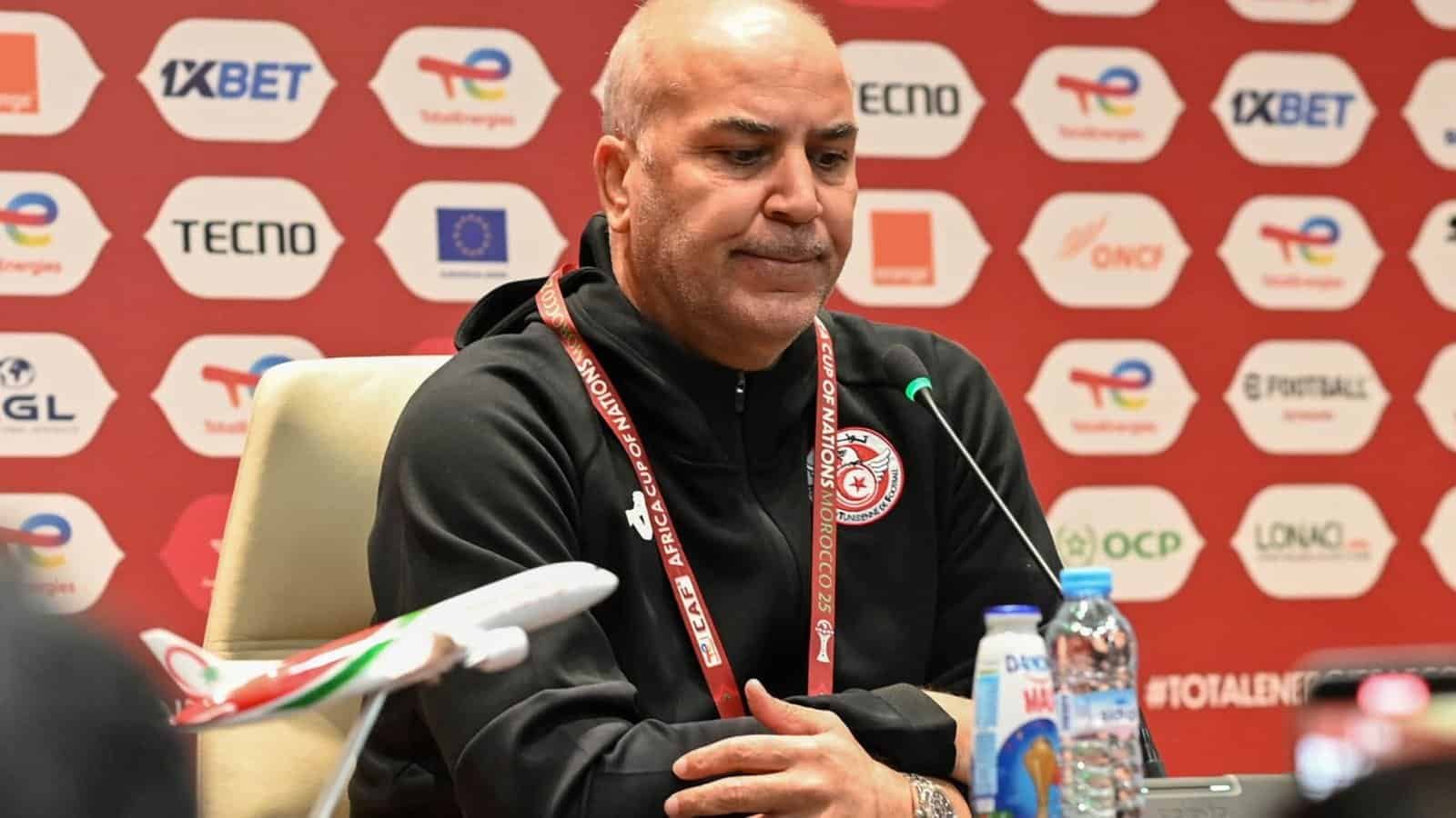 Tunisian coach seated at a press conference with a serious expression, surrounded by branding elements for the Africa Cup of Nations.