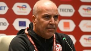 Tunisia's national team coach, focused and serious, speaks at a press conference backdrop featuring sponsorship logos and national colours.